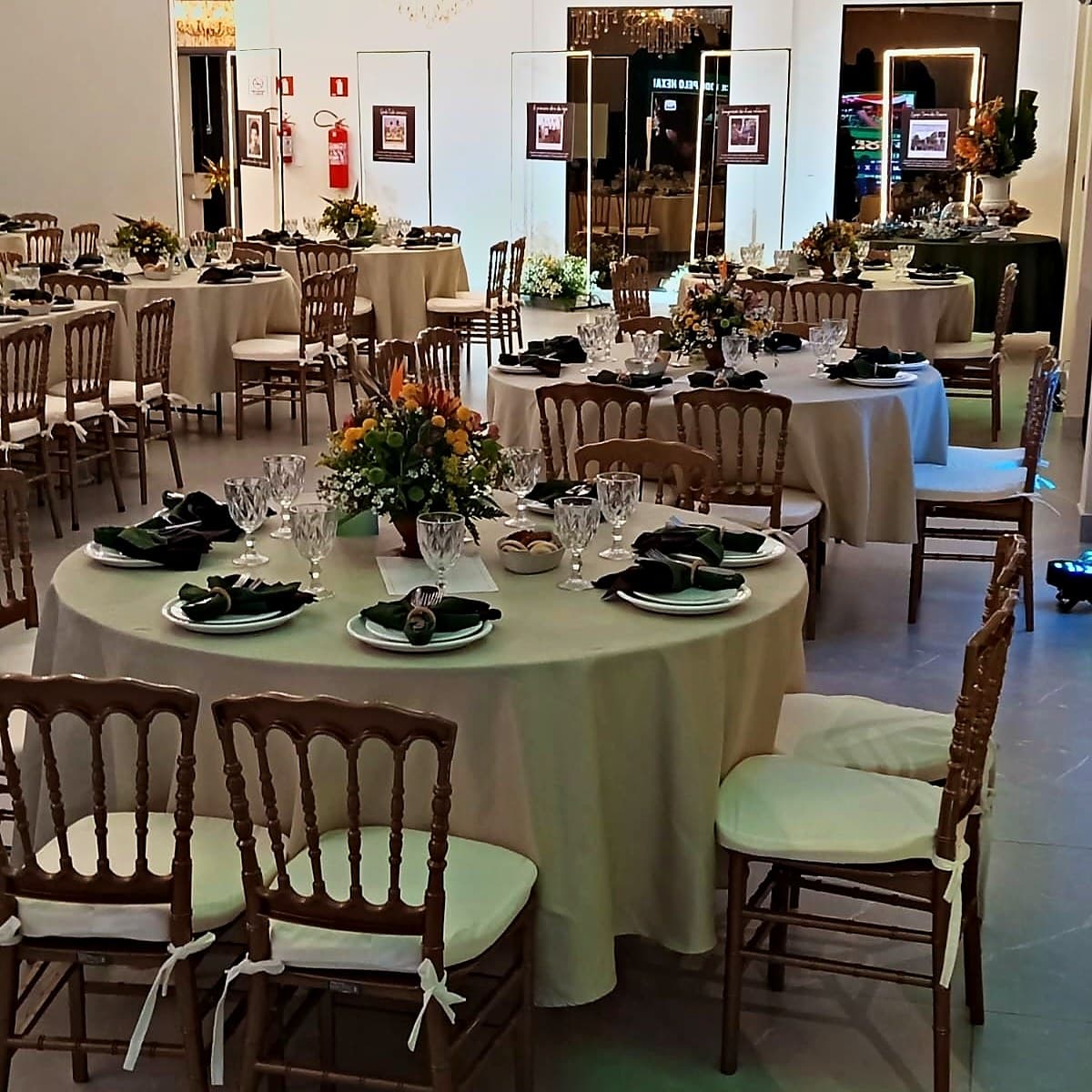 Salão de eventos montado com mesas redondas, arranjos de flores e mesa posta